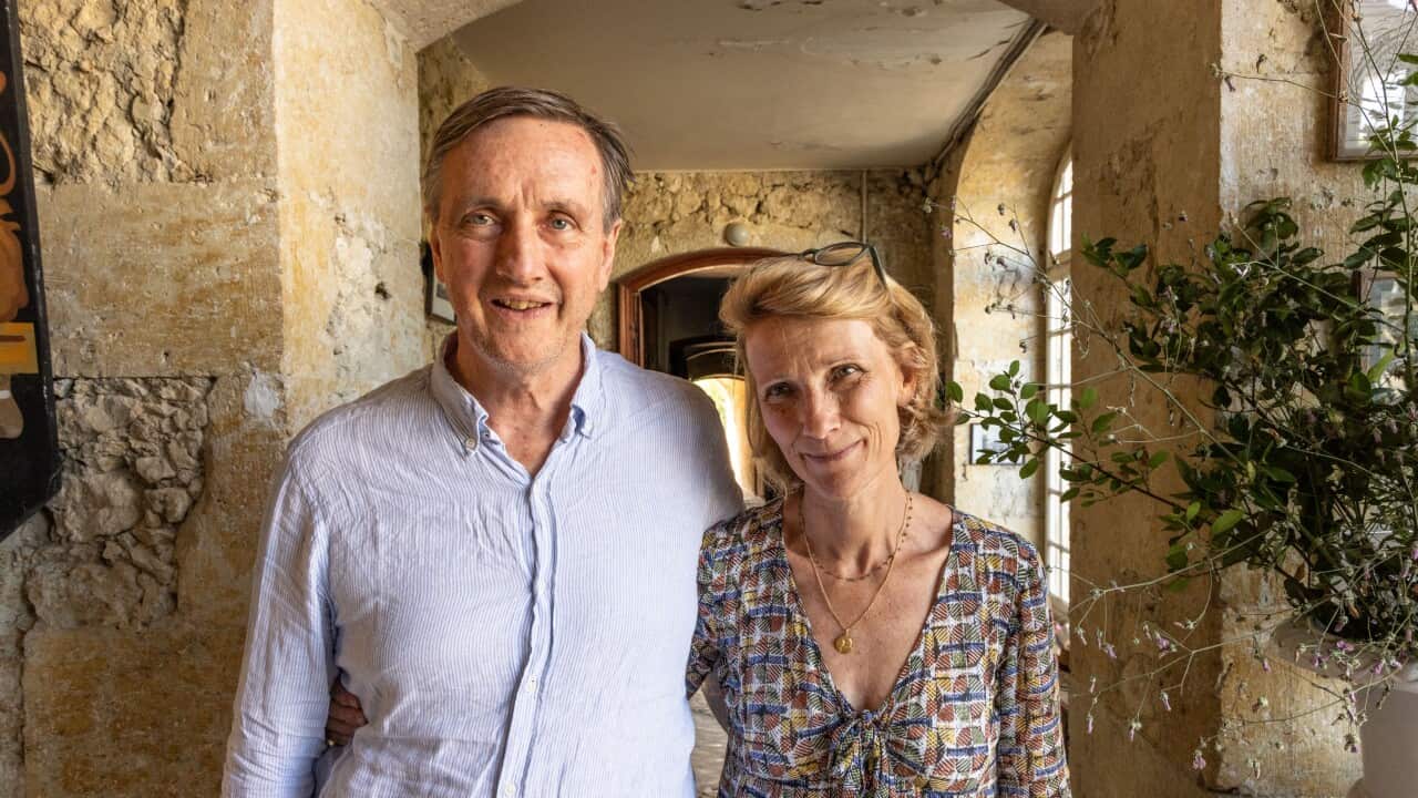 A middle-aged couple with their arms around each other smile. They are standing in the hallway of an old-looking house.