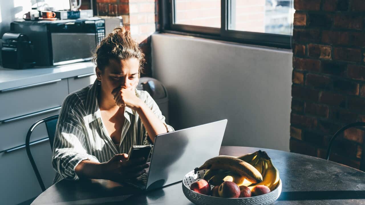 A person sits at a round kitchen table in a sunlit room, looking anxiously at a smartphone while a laptop and a bowl of fruit sit before them.