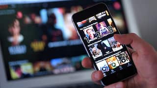 A person holding a phone with the Netlix app open. A laptop in the background also has Netlflix open.