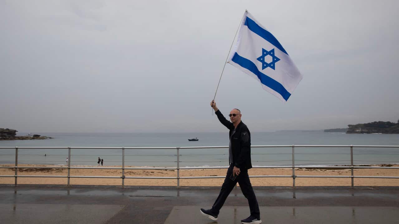 A man holding an Israeli flag as he walks along a promenade, a beach is in the background.