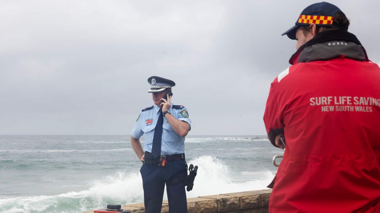 A police officer in a light blue uniform speaks on a mobile phone near a stone wall overlooking a rough ocean, while a person in a red Surf Life Saving New South Wales jacket watches from the foreground.