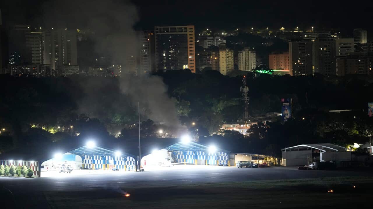 A high-angle nighttime view of an airfield featuring illuminated hangars and a small private jet, with dark smoke rising from behind the hangars against a backdrop of city buildings.