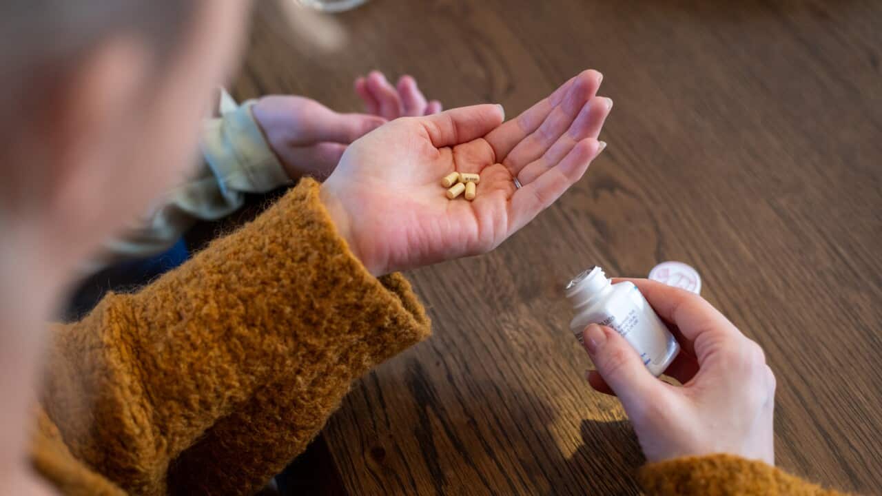 A person in a mustard jumper with pills in one hand and a medication bottle in the other