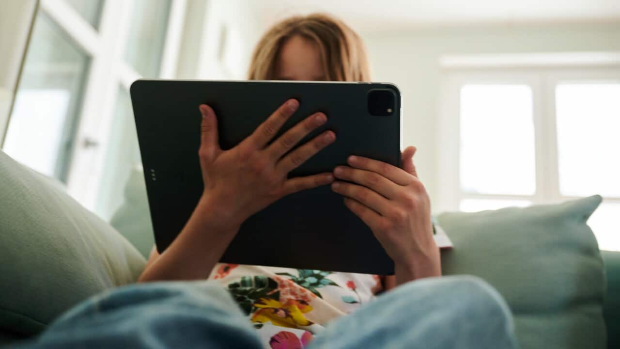 A person with long blonde hair sitting on a couch looks at a large tablet device.