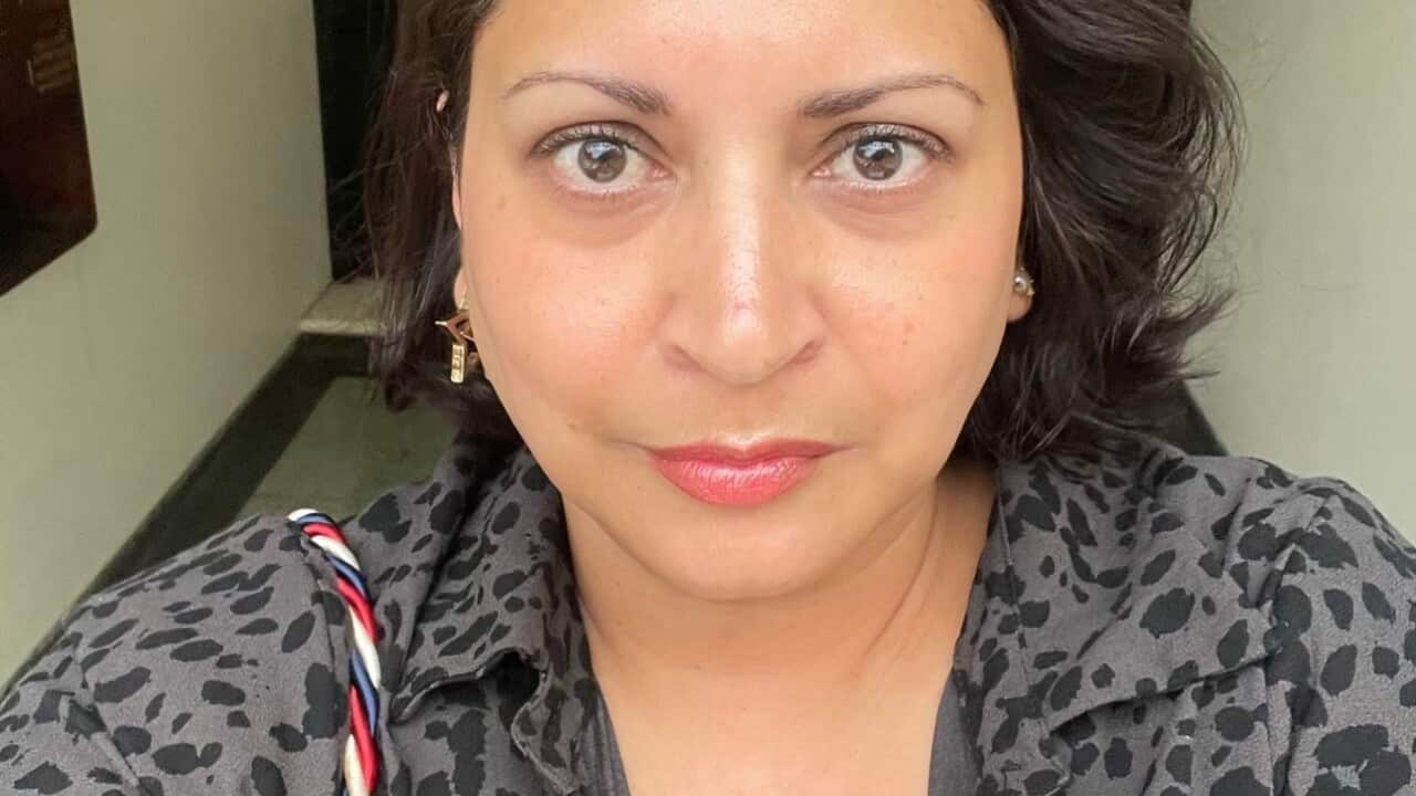 A selfie of a woman with dark hair standing in a hallway with a neutral expression on her face.