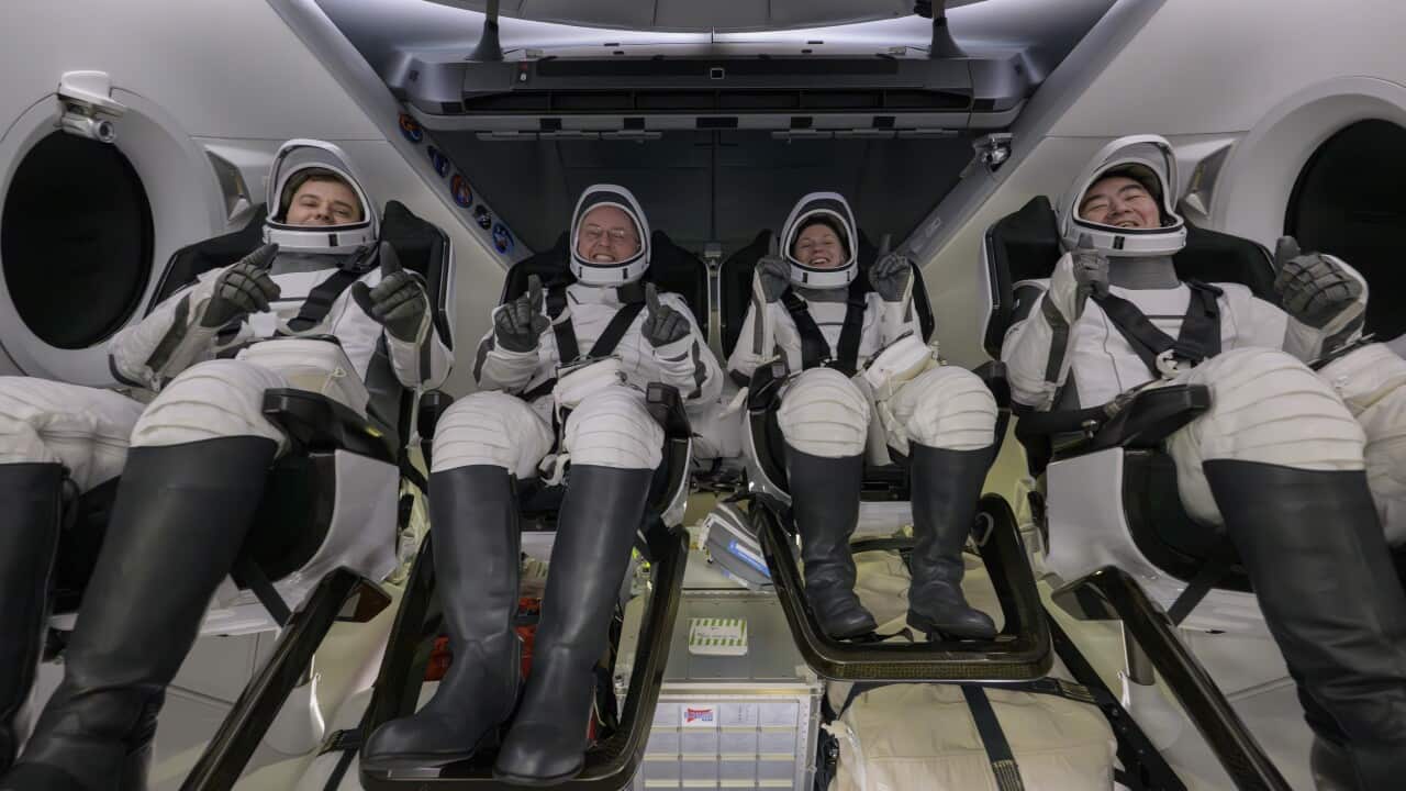 Four astronauts wearing white space suits and seated inside spacecraft chairs. They are giving thumbs up or holding their hands up and smiling.