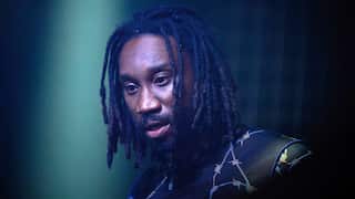A man with dreadlocks in a top with a barbed wire pattern stares ahead, slightly downward.