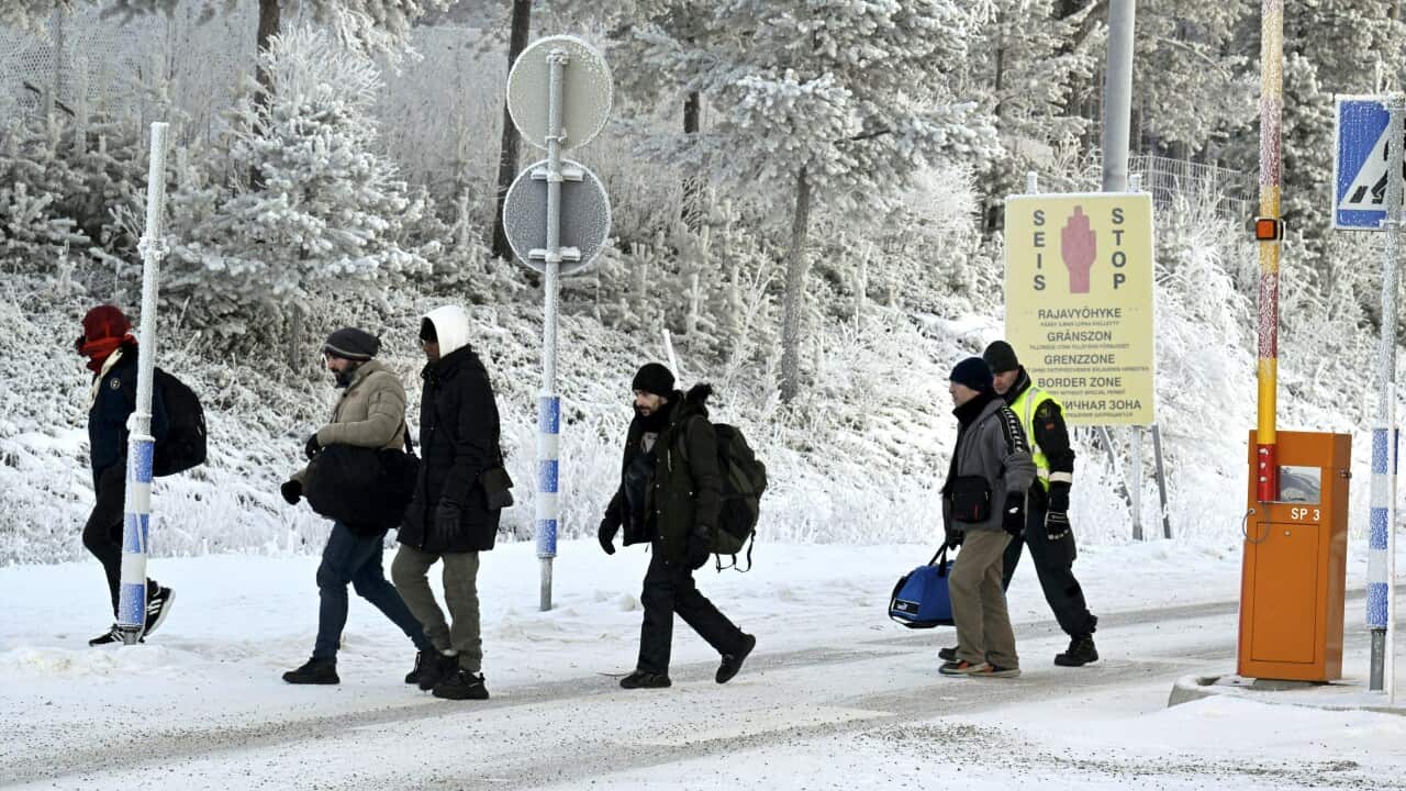 A guard escorts migrants wearing winter clothing across a snow-covered road.