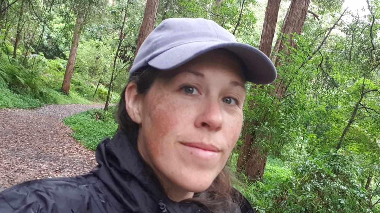 A woman in a forest wearing a black jacket and grey cap
