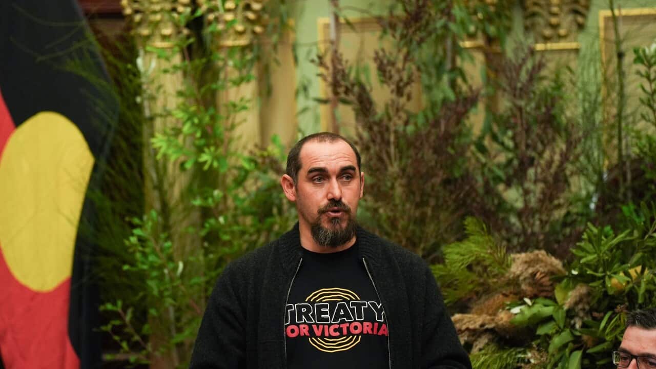 Rueben Berg stands on the stage delivering a speech. An Aboriginal flag can be seen behind him.