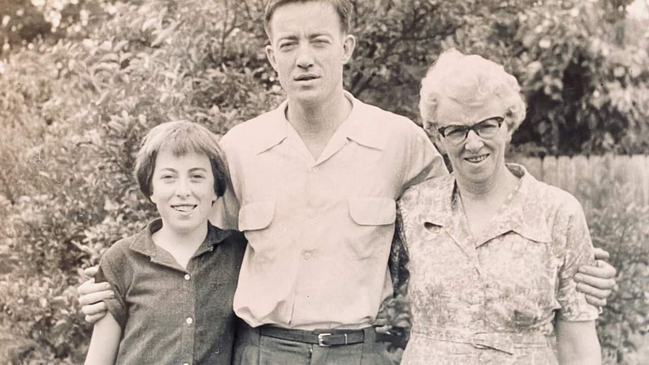 a black and white photo from the 1950s of a tall young man with his arms around a young girl and an older woman, standing in front of a bush