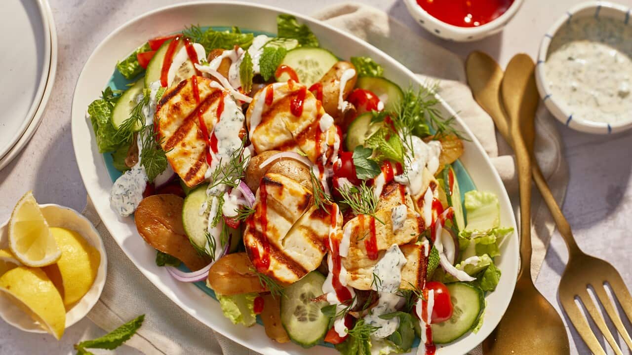 Seen from overhead, an oval platter sits on a surface alongside cutlery, two small bowls with dressing and another bowl with lemon wedges. The platter holds a salad topped with grill-marked slices of haloumi and drizzled with a red dressing and a creamy dressing.
