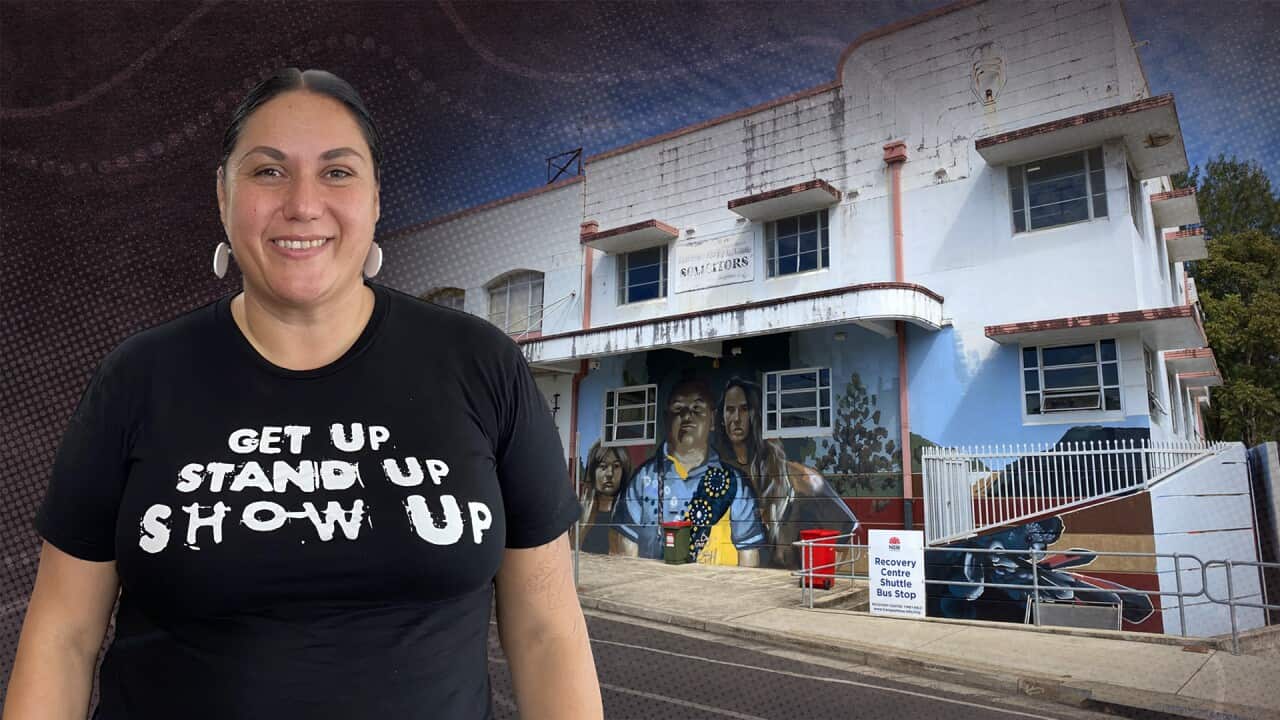 a composite of a smiling woman wearing a black tshirt with the words "get up stand up show up" in white, superimposed over a street shot of a blue two story mid-century building exterior that has a mural drawn over its lower level.