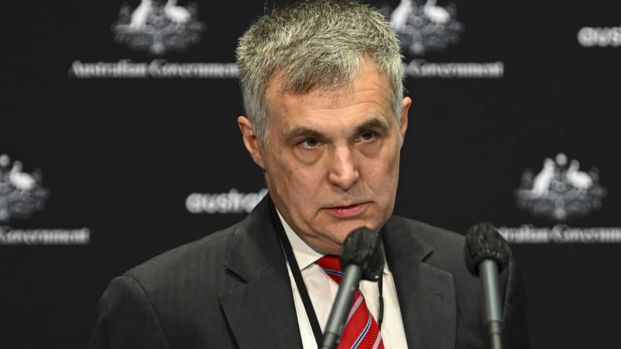 A medium close-up photograph shows a middle-aged man with grey hair wearing a dark suit, white shirt, and a red-and-white striped tie. He stands behind a microphone in front of a dark backdrop with repeating "Australian Government" and Coat of Arms logos.