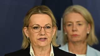 A woman with blonde hair in a dark suit stands in front of two other people