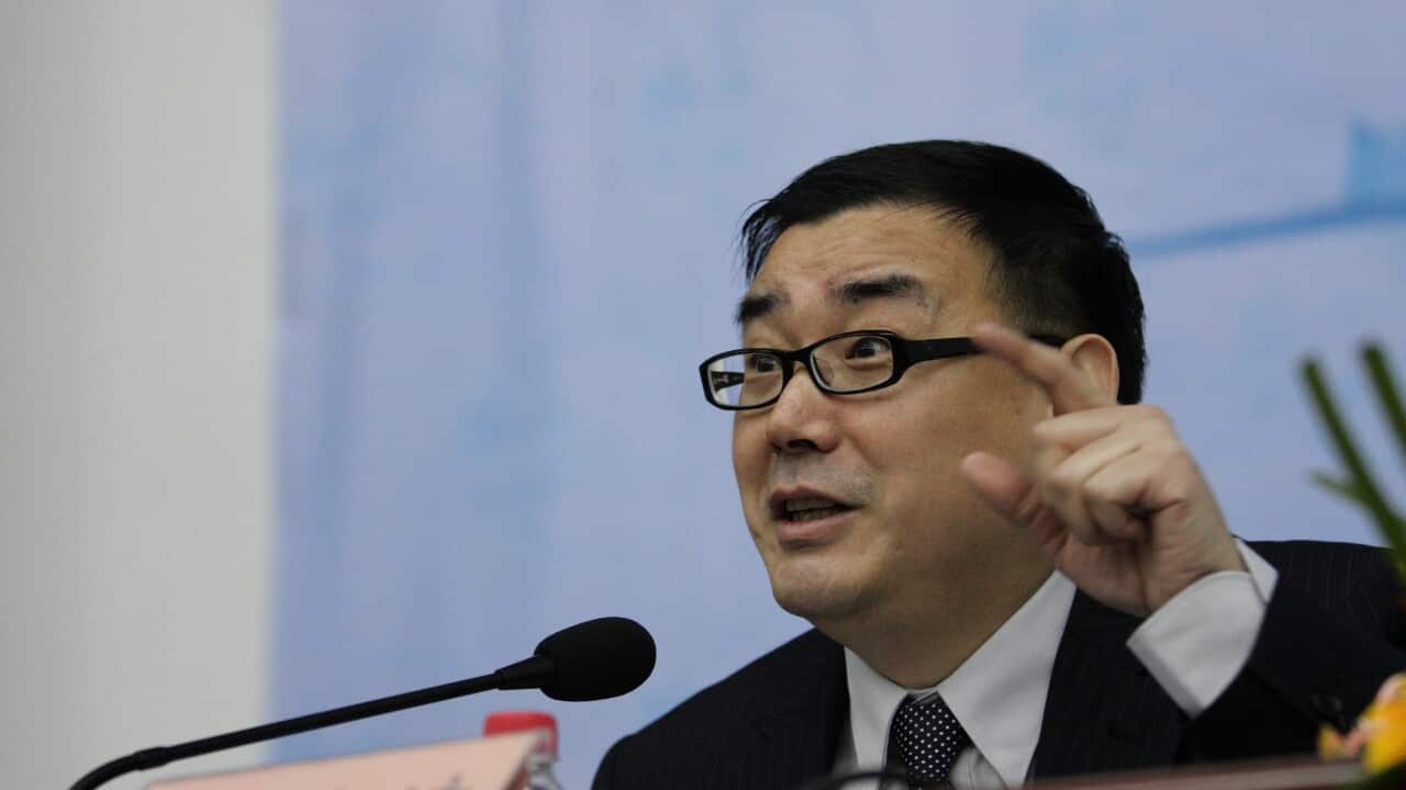 A man of Asian descent wearing glasses is speaking into a microphone with his hand raised.