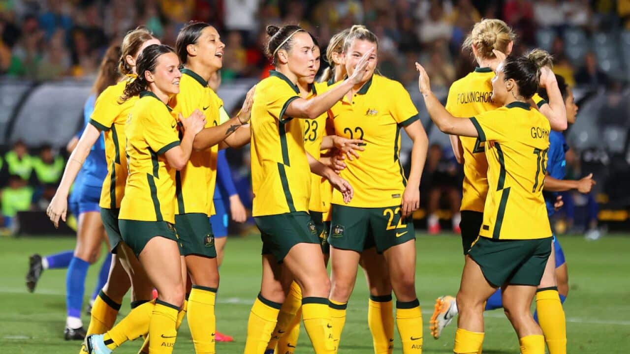 Women footballers wearing yellow jerseys, green shorts and yellow socks celebrate a goal