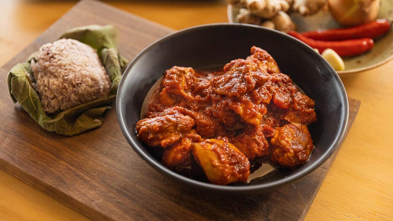 A dark bowl sits on a woooden board, on a yellowish surface. The bowl holds a pieces of chicken in a thick red sauce. Leaf-wrapped rice sits alongside.