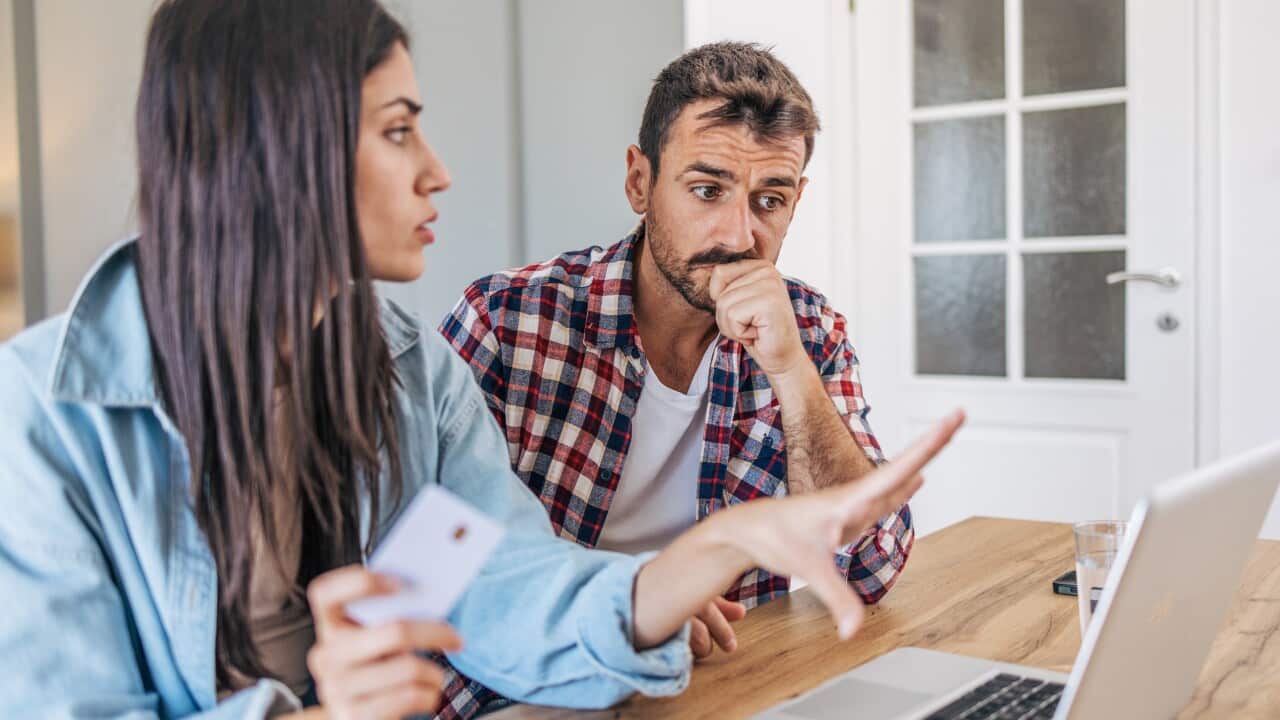 A couple sitting at the table, using a laptop, with the living room visible in the background. They are shopping online with a credit card and look concerned.