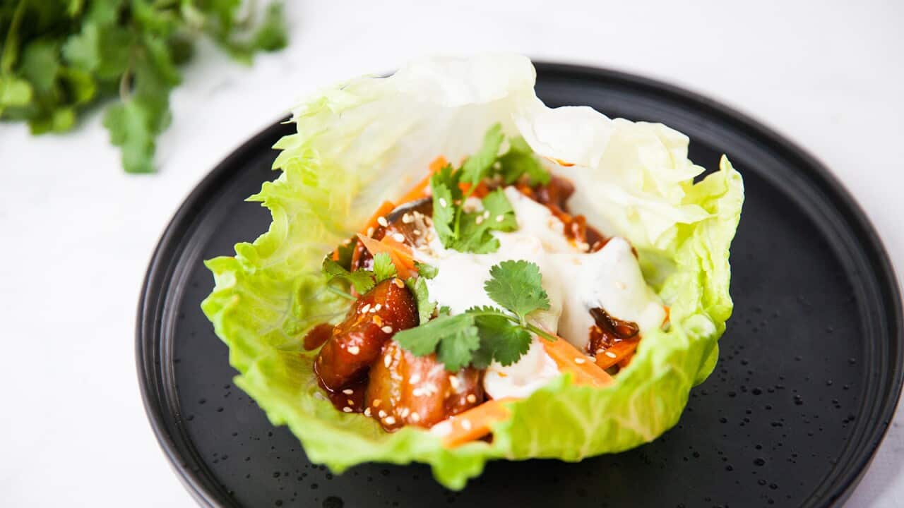 A vibrant green iceberg lettuce leaf sits on a flat black plate. The lettuce cup holds sticky brown eggplant pieces and carrot matchsticks, drizzled with a creamy sauce and garnished with fresh coriander sprigs.