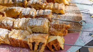 Five metal skewars, threaded with toasted brioche cubes, sit on a round wood and pink platter. The kebabs have been drizzled with a creamy-yellowish sauce and dusted with icing sugar.