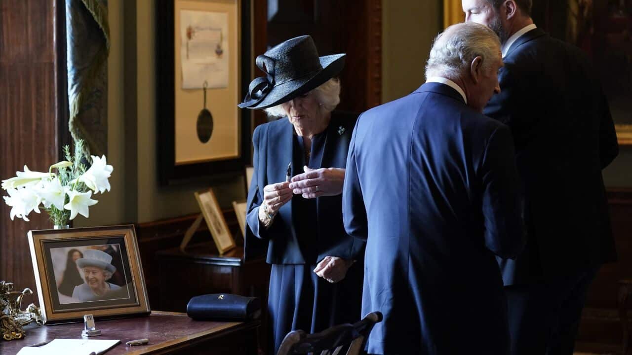 Queen Consort Camilla holds a pen with King Charles III and another man standing next to her.