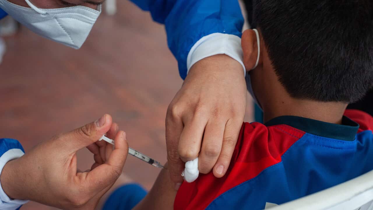 A nurse wearing scrubs and a surgical mask inserts a syringe into a child's shoulder