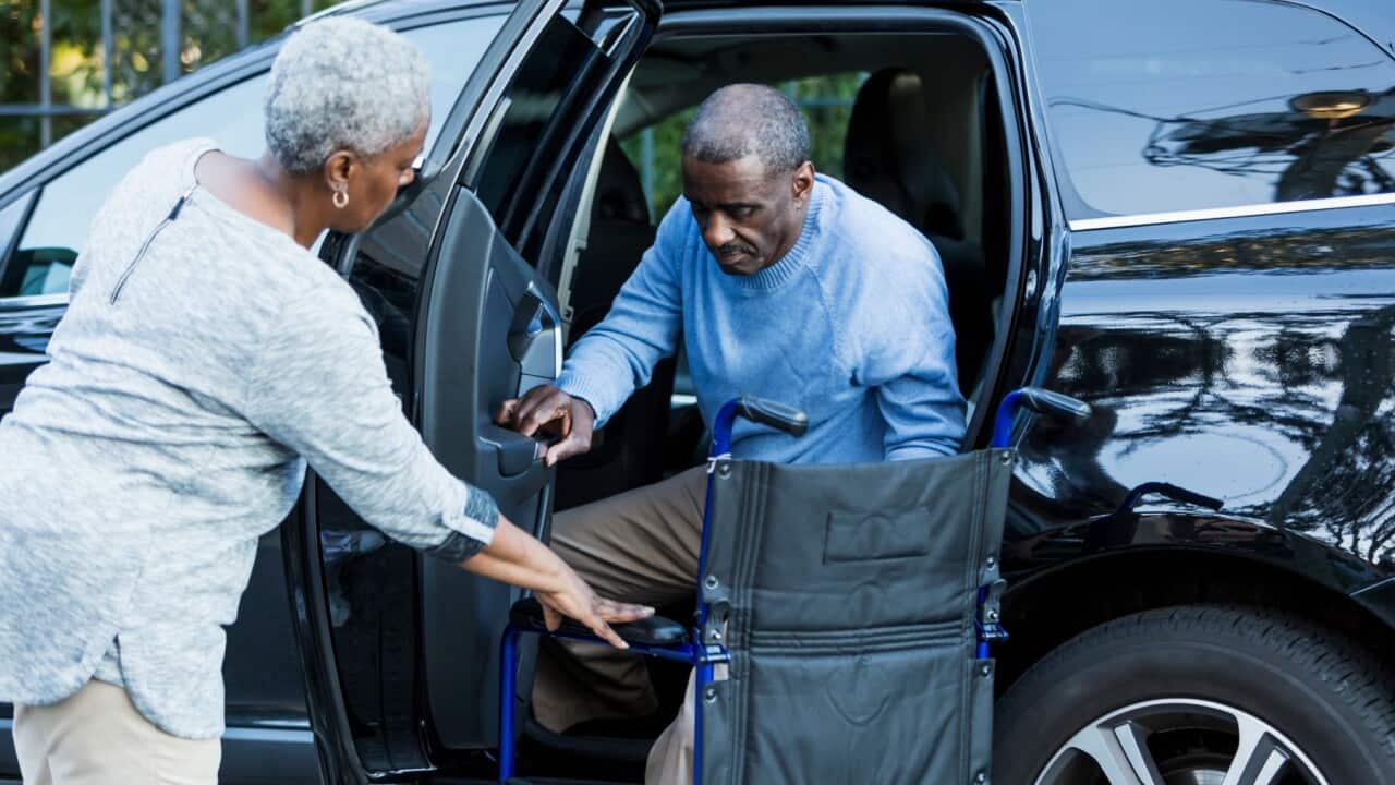 A senior African American couple parked in a handicapped parking space. The man is getting out of the car and his wife is helping him into his wheelchair.