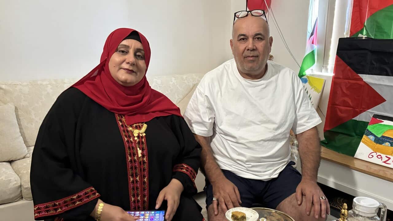 A couple is seated in a living room, with a woman in a black and red embroidered dress and hijab holding a phone, next to a man in a white t-shirt and dark shorts. They are positioned behind a large platter of baklava and other sweets, with a Palestinian flag visible in the background.