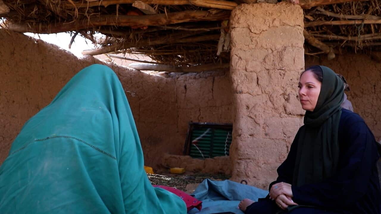 A woman covered in a blue robe talks to a woman in a black shawl. Both are sitting on the ground