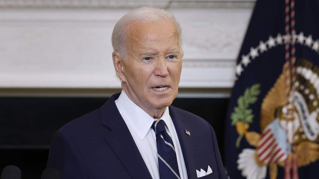 A man in a dark suit with white hair stands in front of a lectern. Behind him hangs a flag bearing the US coat of arms.