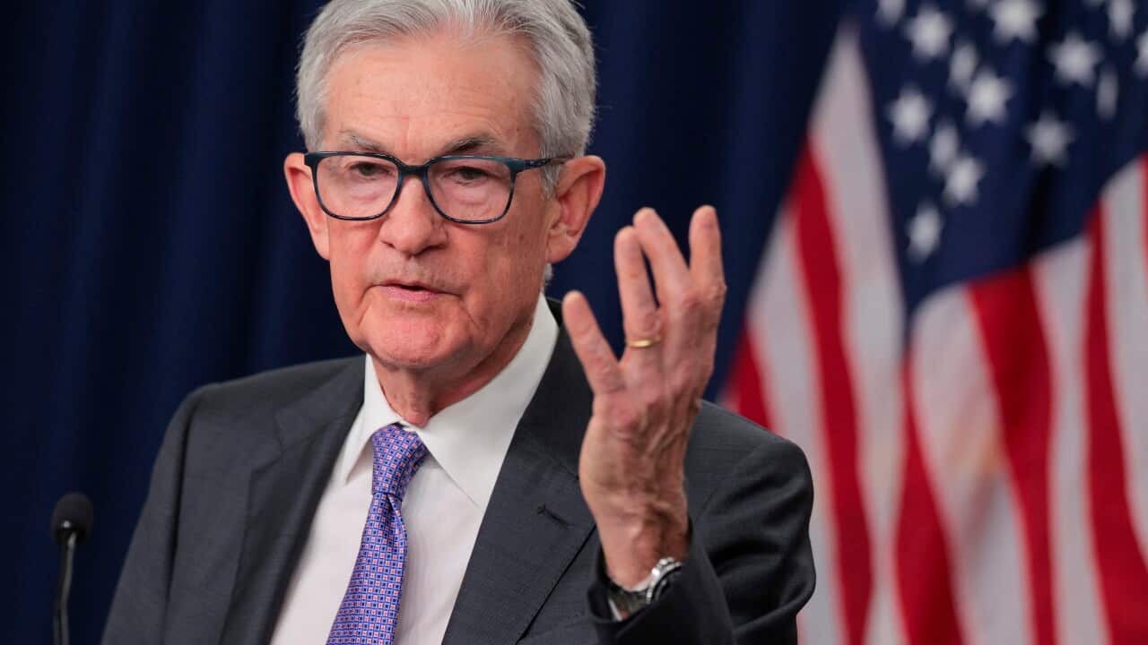 An elderly man in a suit raises his hand as he speaks at a lectern.