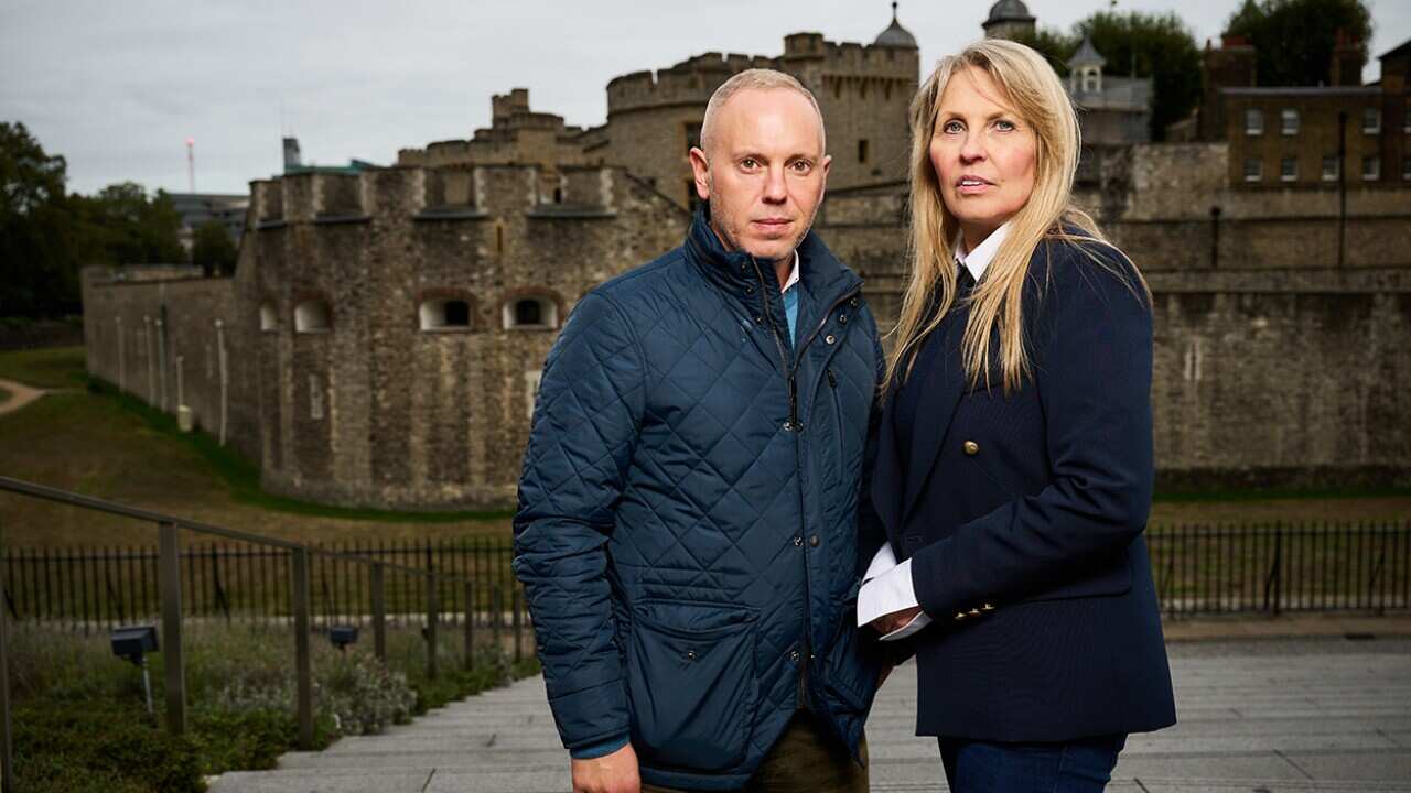 A man and a woman stand in front of a grey historic stone building.