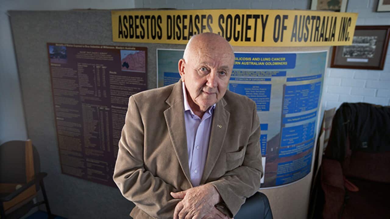 Balding elderly white man wearing brown jacket leans on office chair with "Asbestos Diseases Society of Australia" sign behind him.