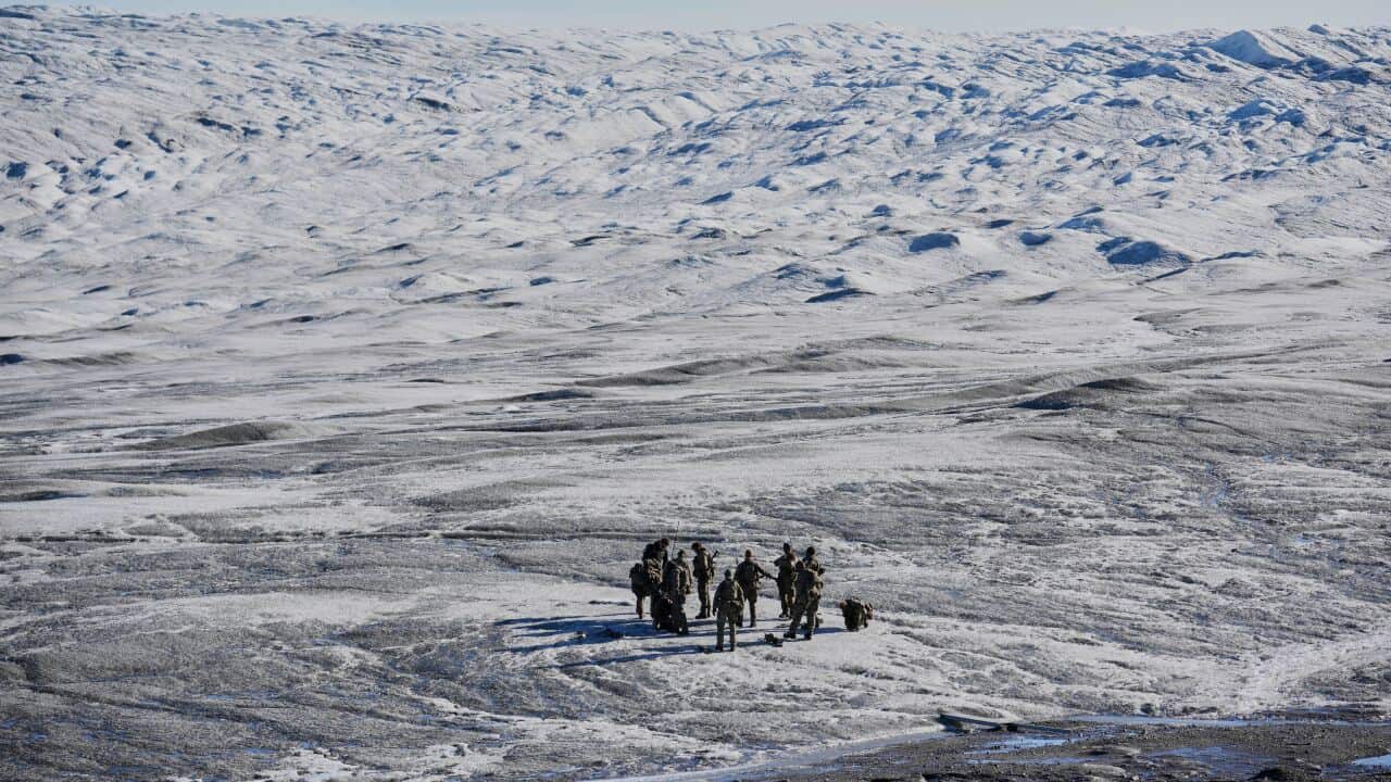 A group of military personnel stands in a huddle on a vast, rugged, and snow-dusted landscape under a clear sky.