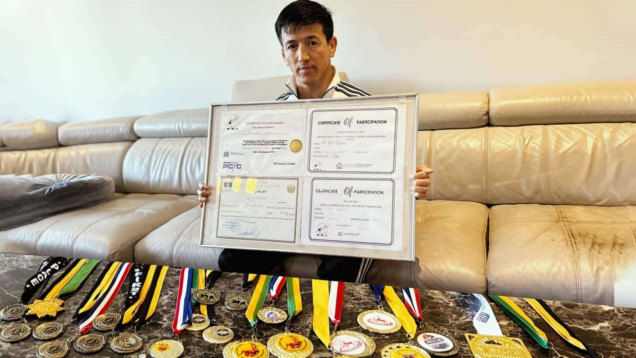 A man holds up a frame of certificates as many medals are displayed on a table in front of him