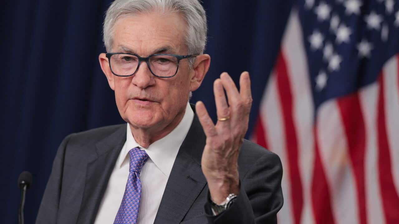An elderly man in a suit raises his hand as he speaks at a lectern.
