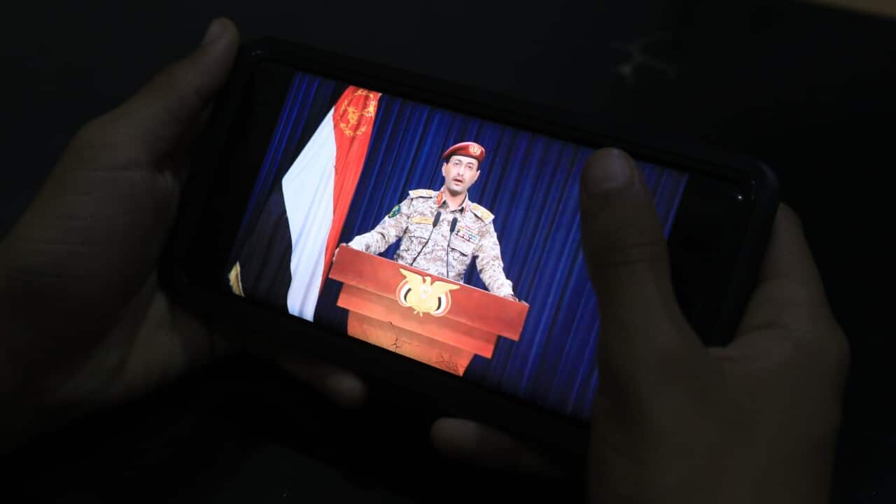 A person watches on a mobile phone a man in military uniform speaking from behind a lectern.
