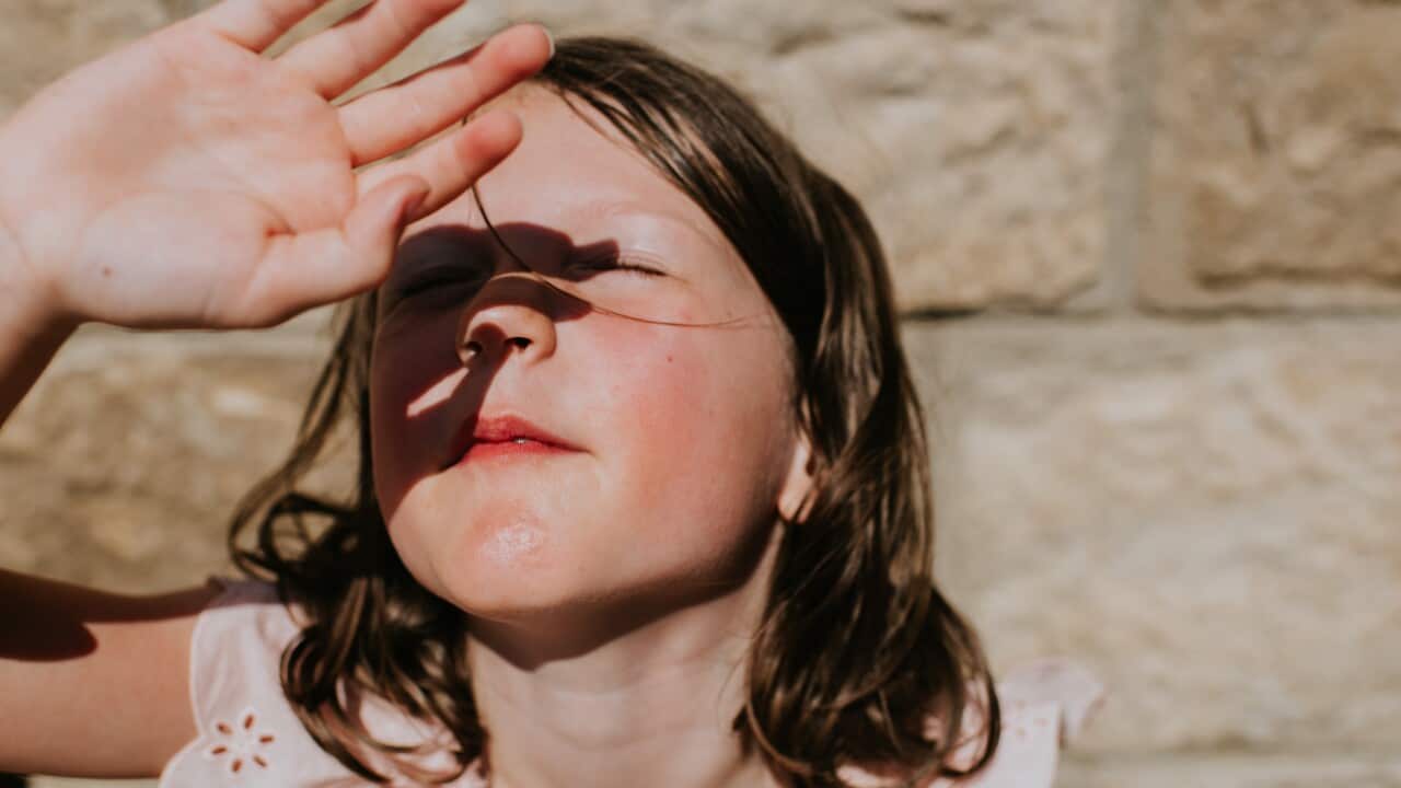 A little girl raises her hand and covers her eyes to protect them from the bright sunlight. She smiles.