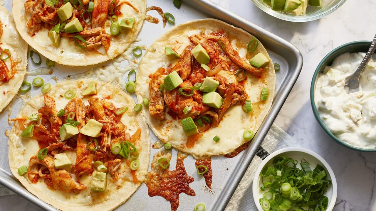 A metal baking tray sits on a marble surface. Three open tortillas, and a little of a fourth, sit on the tray, each topped with a saucy chicken mix and cubes of avocado. Bowls with sour cream, avocado and sliced green onion sit alongside the tray.