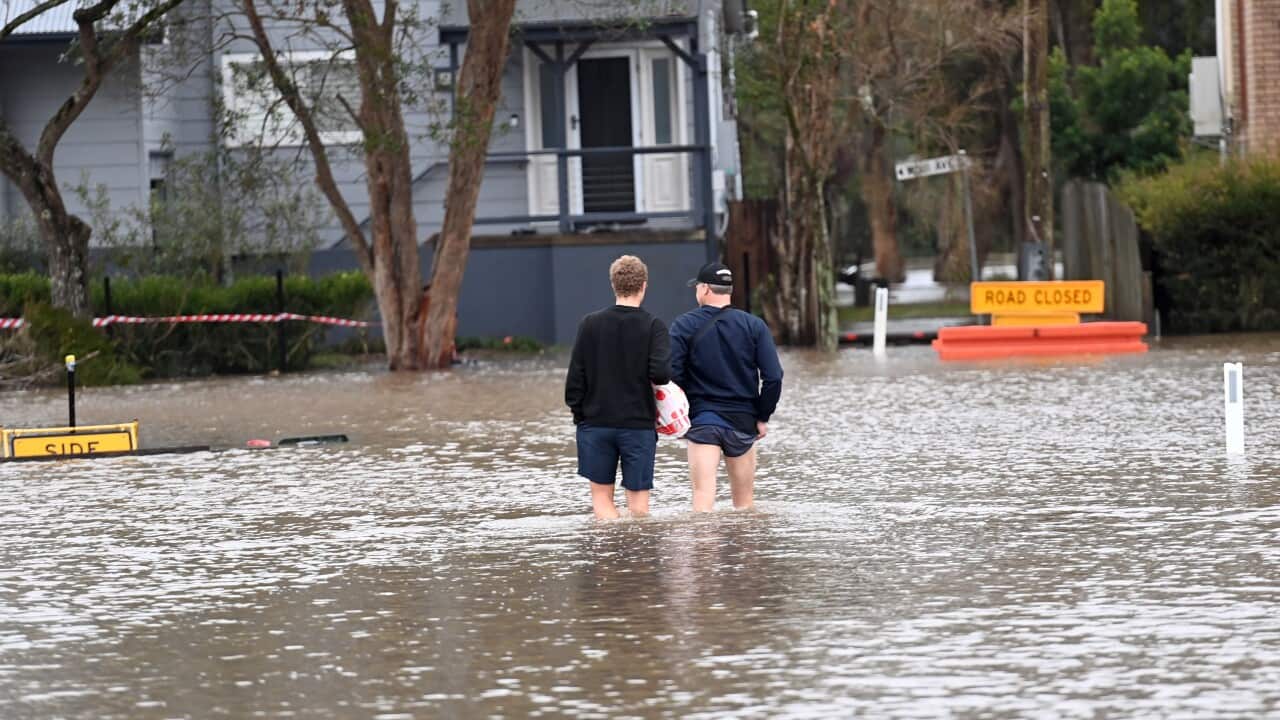 Two men facing away from camera wade through floodwaters toward house.