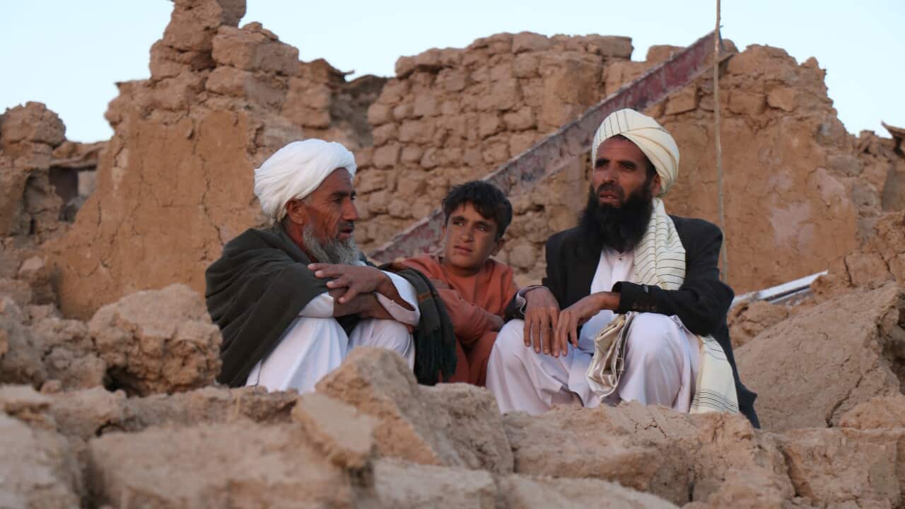 Two bearded older men wearing traditional Afghani clothing and a younger boy sit in the rubble of a building.