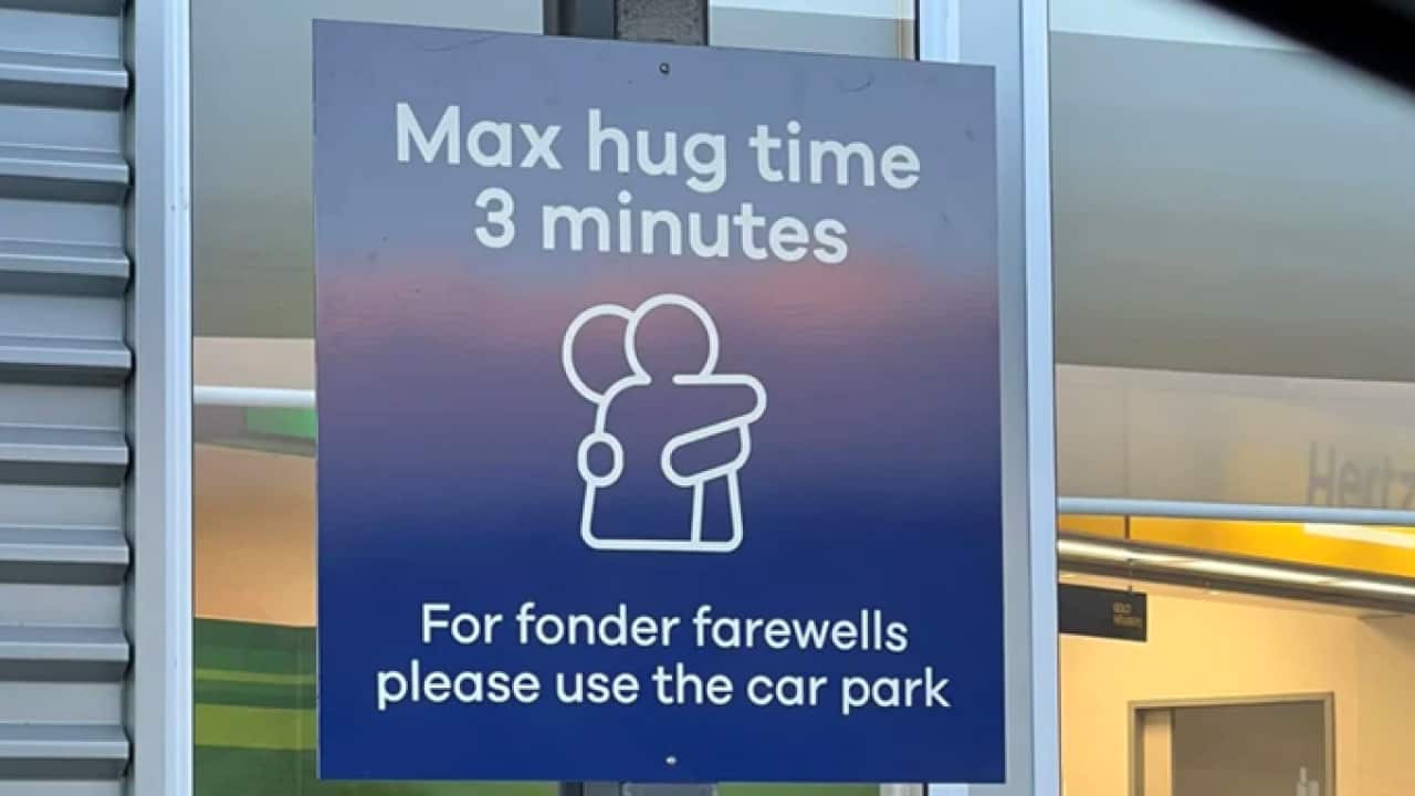 A blue sign features 'Max hug time 3 minutes' at the top and a message at the bottom stating, 'For longer farewells, please use the car park.' In the centre, there is an image of two people hugging.
