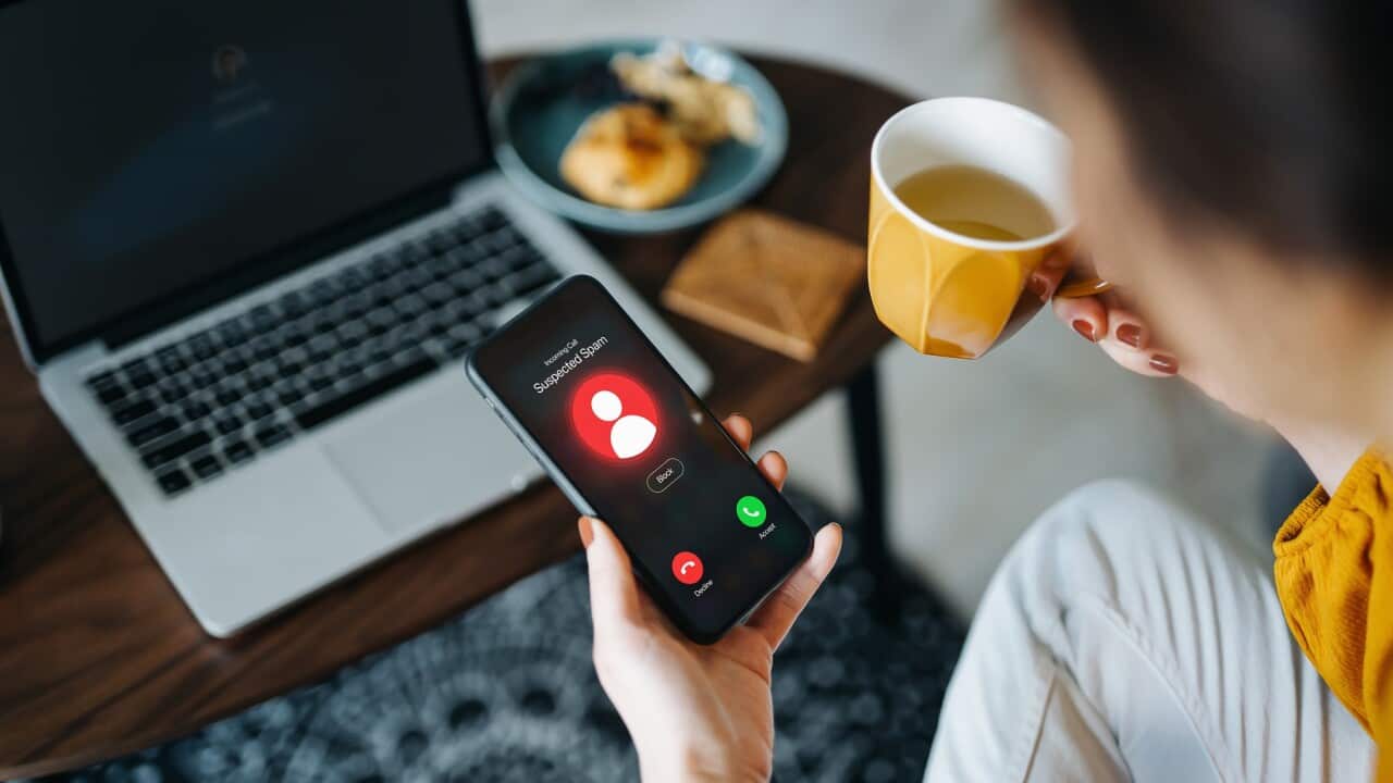 Over the shoulder view of young Asian woman receiving an incoming suspected call from unknown caller on her smartphone and rejecting the call at home. Device screen showing warning sign as detected by the network provider. Phone scam and fraud concept