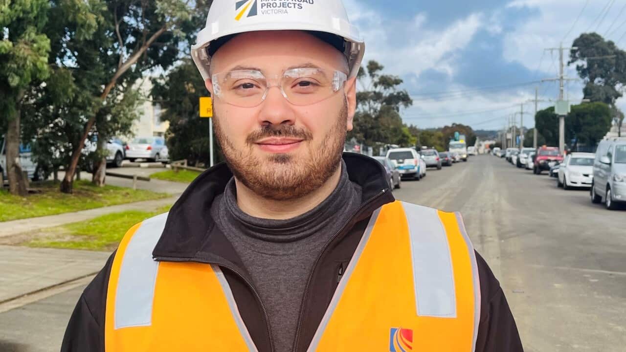 A man wearing an orange hi-vis vest, dark sunglasses and a white hard hat