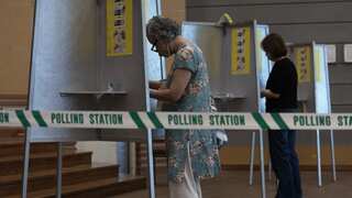 Two pepole voting at a polling station. There is a line of green tape that says "polling station" in front of them.
