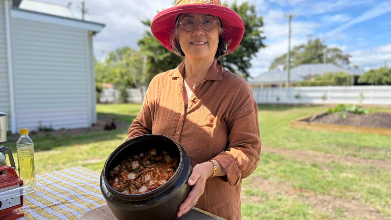 Woman in an orange hat and orange shirt smiles and holds a black pot full of kimchi