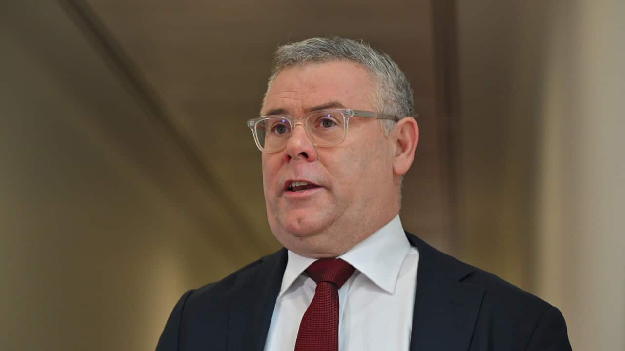 A man wearing a dark blue blazer, white shirt and red tie. He is wearing clear glasses and has short grey hair.