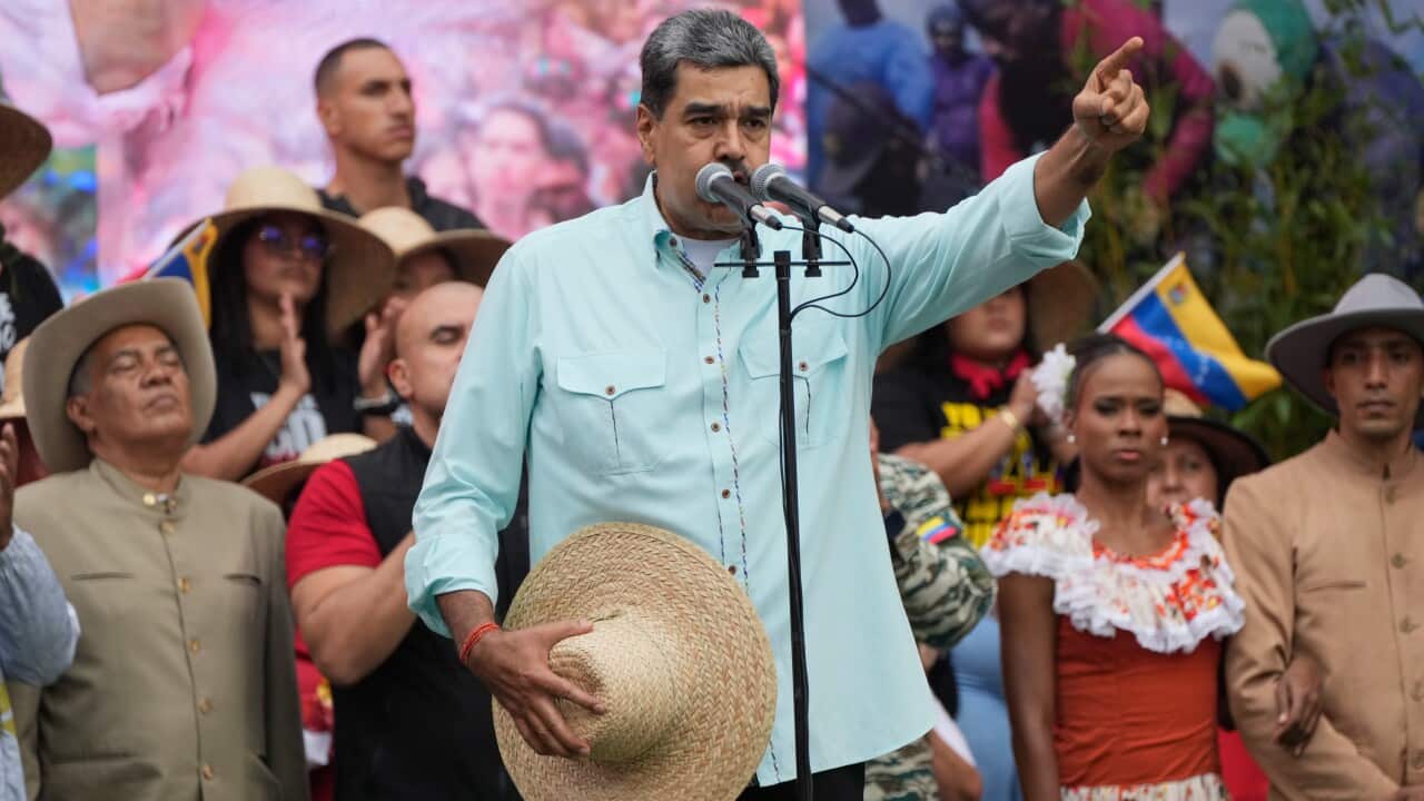Venezuelan President Nicolás Maduro gestures toward a crowd while speaking at a podium, holding a straw hat in front of supporters dressed in traditional and casual attire.