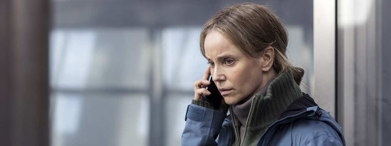 A woman in a heavy blue jacket stands in front of a grey-toned window, talking on a mobile phone.